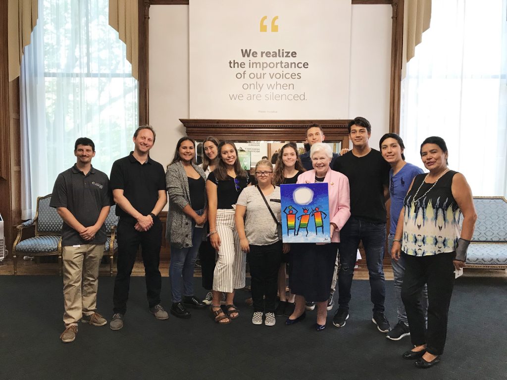 Youth participants of Nikibii Dawadinna Giigwag program stand with Her Honour in Queen's Park with a paining of three Indigenous figures made for Her Honour