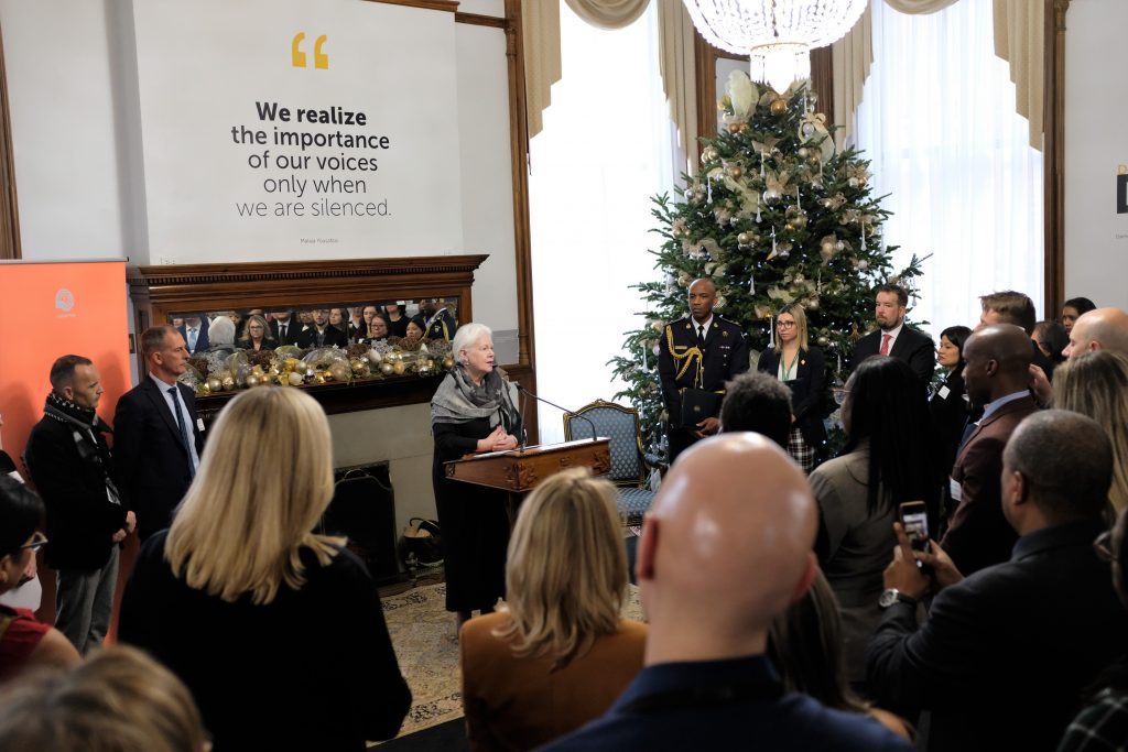 The Lieutenant Governor stands a podium giving remarks to a large grouping of Ontario Public Servants who are facing her. A large Christmas tree is to her right.