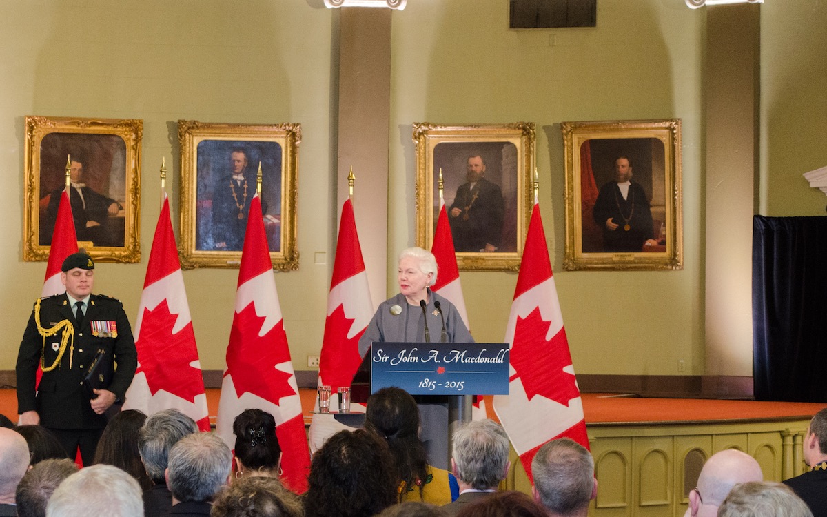 The Lieutenant Governor stands at a podium with five Canadian flags standing behind her and beyond that gold framed painted portraits of Kingston leaders