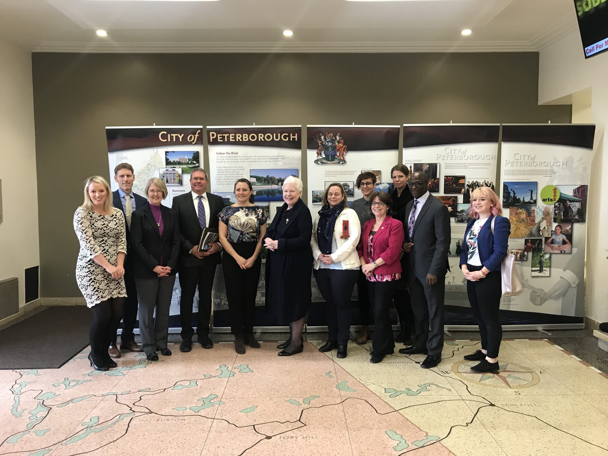 The Lieutenant Governor stands with the Mayor of Peterborough Diane Therrien along with staff in the foyer of Peterborough City Hall. The floor is a large map of the region.